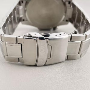 May include: Close-up of a silver Bulova watch clasp. The clasp is made of polished stainless steel and features the Bulova logo. The watch band is also silver and made of stainless steel. The watch face is visible in the background.