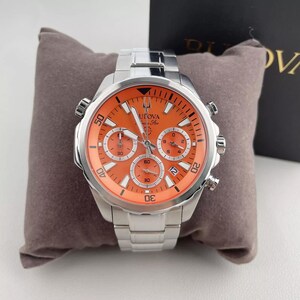 May include: A stainless steel Bulova watch with an orange face and silver band. The watch features three sub-dials and a date window. The watch is displayed on a gray pillow.