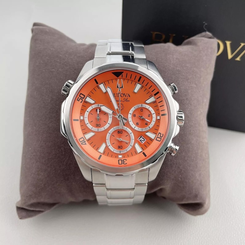 May include: A stainless steel Bulova watch with an orange face and silver band. The watch features three sub-dials and a date window. The watch is displayed on a gray pillow.