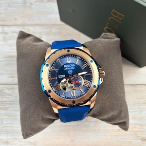 Pu&ograve; includere: Un orologio automatico Bulova Marine Star con un design blu e oro. L'orologio presenta un quadrante blu con numeri romani, un movimento a vista e un cinturino in gomma blu. L'orologio &egrave; esposto su un cuscino grigio.