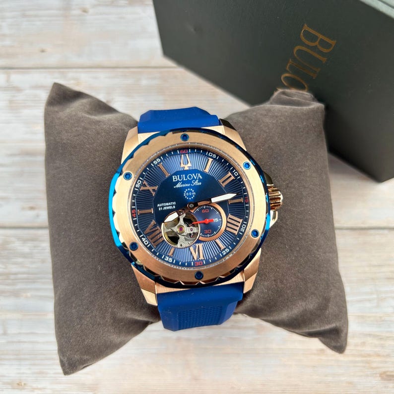 Pu&ograve; includere: Un orologio automatico Bulova Marine Star con un design blu e oro. L'orologio presenta un quadrante blu con numeri romani, un movimento a vista e un cinturino in gomma blu. L'orologio &egrave; esposto su un cuscino grigio.