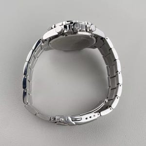 May include: A silver-toned stainless steel chronograph wristwatch featuring a polished link bracelet, a round case, and a side crown embossed with a logo. The watch includes two side pushers and a secure fold-over clasp mechanism.