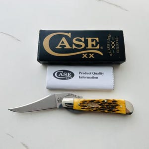 May include: A Case XX pocket knife with a curved blade and a yellow and brown handle. The knife is next to its black box, which has the Case XX logo and text. A white paper with the Case XX logo and "Product Quality Information" is also present.