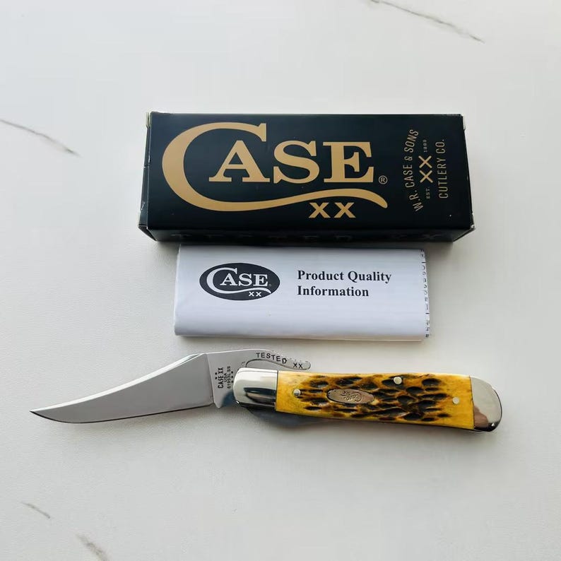 May include: A Case XX pocket knife with a curved blade and a yellow and brown handle. The knife is next to its black box, which has the Case XX logo and text. A white paper with the Case XX logo and "Product Quality Information" is also present.