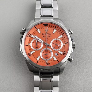 May include: A stainless steel Bulova Marine Star chronograph watch with a vibrant orange dial, silver-tone hands, and hour markers. The watch features three sub-dials, a date window at the four o'clock position, and a metallic link bracelet.