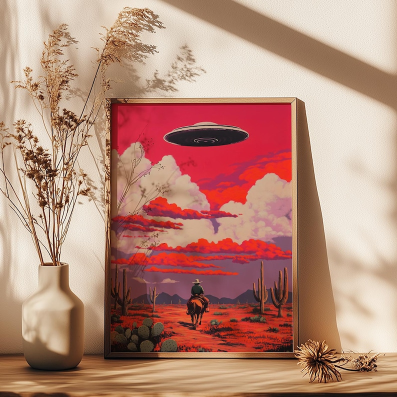 May include: A framed illustration of a cowboy riding a horse in a desert landscape with a UFO flying overhead. The sky is a vibrant pink and orange with fluffy white clouds. The desert is a reddish-brown color with cacti and a few trees.