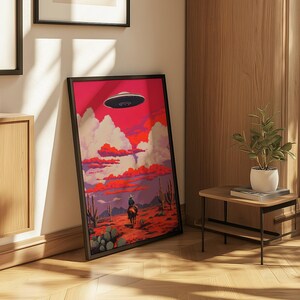 May include: A framed print of a cowboy riding a horse in a desert landscape with a flying saucer in the sky. The sky is a vibrant pink and orange with fluffy white clouds. The print is framed in black.