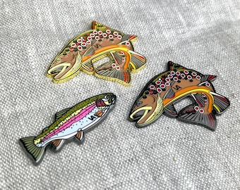 May include: Three enamel pins in the shape of fish. Two are brown with orange and red accents, and one is a rainbow trout with pink, green, and white details. The pins are on a textured gray surface.