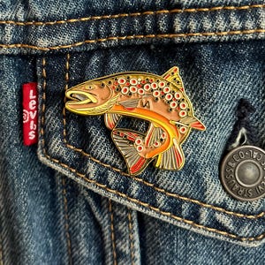 May include: A gold-coloured enamel pin of a brown trout with red and white spots. The pin is on a blue denim jacket.