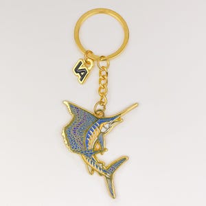 Puede incluir: Un llavero de metal dorado con un dije de pez vela con detalles en esmalte azul y amarillo. El dije está unido a una anilla dorada mediante una cadena corta e incluye un pequeño colgante con las letras VA en color blanco y negro.