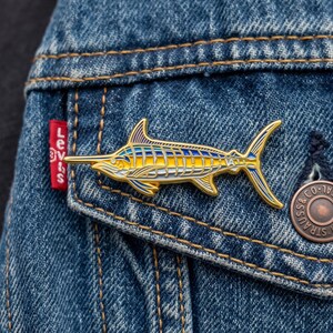 Könnte beinhalten: Eine goldfarbene Emaille-Anstecknadel in Form eines Marlin-Fisches mit leuchtend blauen und gelben Streifen am Körper, einem langen, spitzen Schnabel und detaillierten Flossen, befestigt an der Taschenklappe einer blauen Jeansjacke mit rotem Etikett.