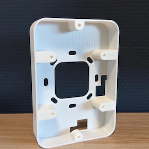May include: A white rectangular mounting bracket with rounded corners, featuring a central square cutout, multiple smaller oval and rectangular openings, and five protruding screw hole tabs positioned along the inner perimeter of the frame.
