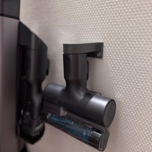 Op de afbeelding: Een zwarte stofzuigeropzetstuk bevestigd aan een muur. Het opzetstuk heeft een cilindrisch ontwerp met een transparant gedeelte dat een blauw onderdeel onthult. De muurbevestiging is ook zwart en lijkt van plastic te zijn.