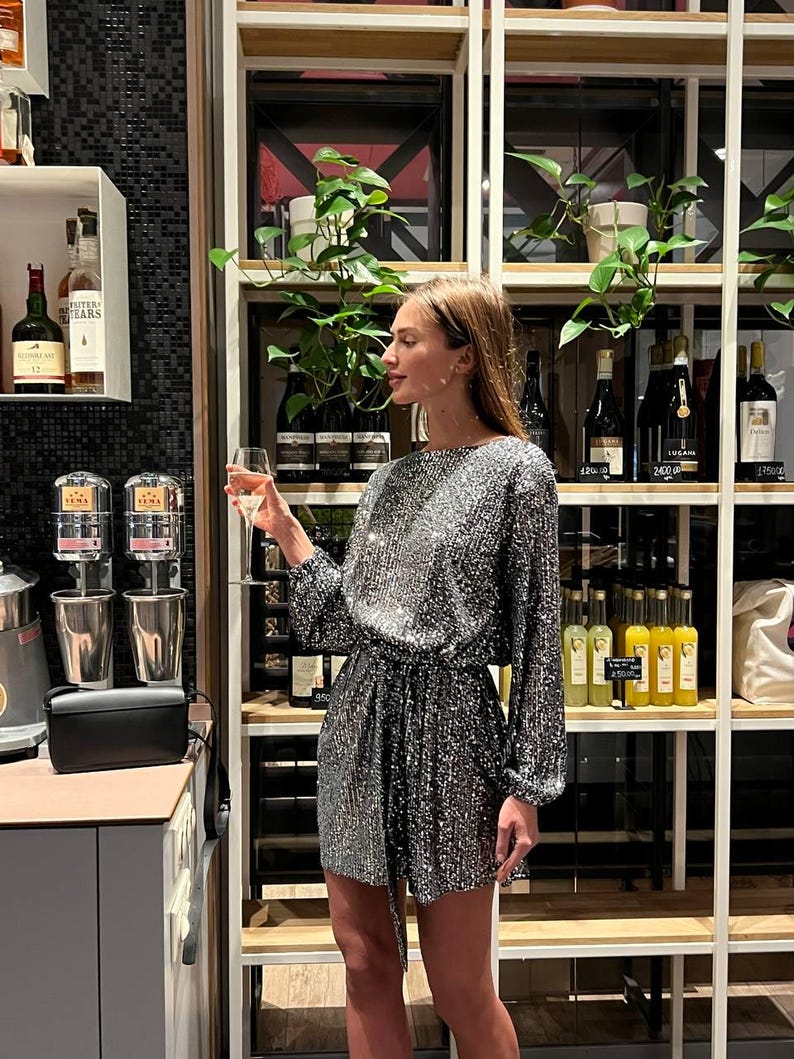 May include: A woman wearing a silver sequined mini dress with a black belt. She is holding a glass of champagne and standing in front of a shelf with wine bottles.