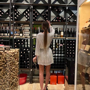 May include: A woman wearing a silver sequined dress and black heels stands in front of a wine cellar. The cellar has shelves filled with wine bottles and a wall of cork stoppers.