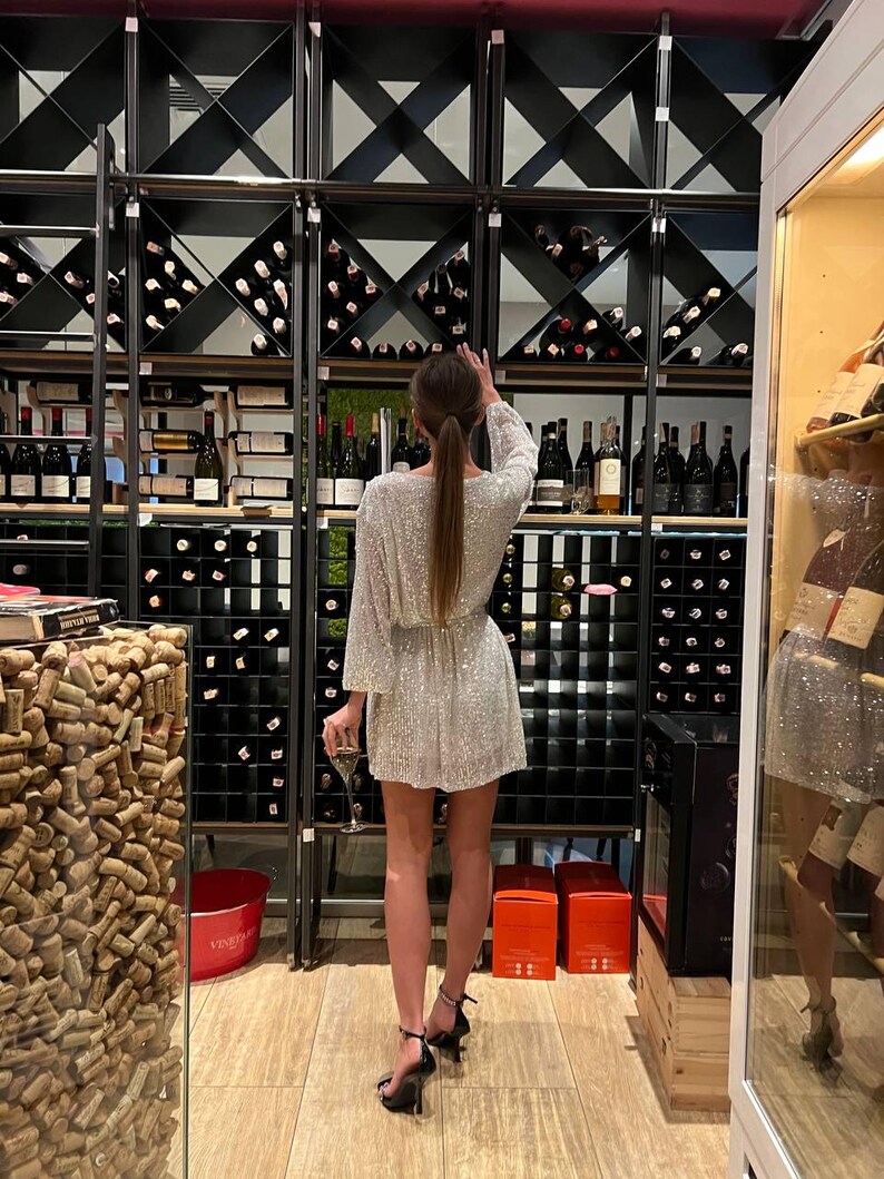 May include: A woman wearing a silver sequined dress and black heels stands in front of a wine cellar. The cellar has shelves filled with wine bottles and a wall of cork stoppers.