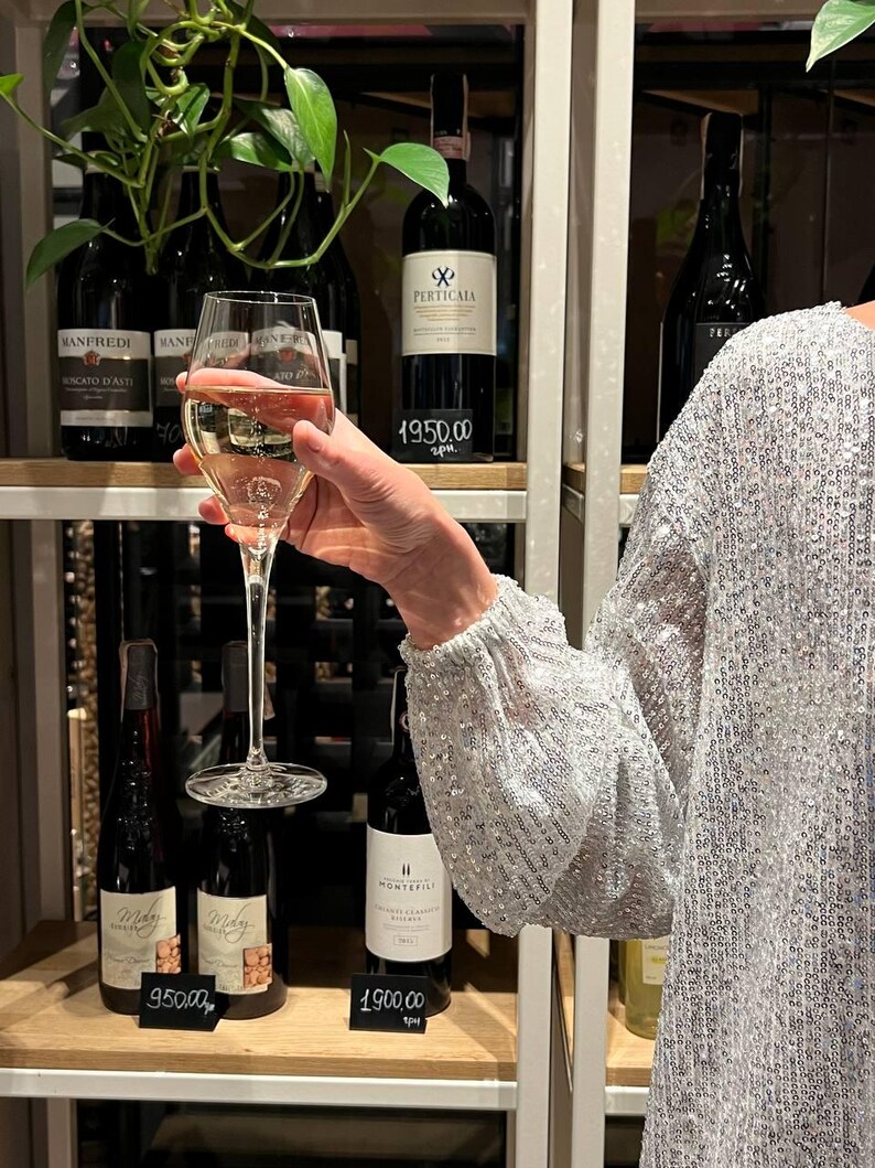 May include: A glass of white wine with a clear stem and a round bowl. The wine is a pale yellow color. The glass is being held by a person's hand. The glass is in front of a shelf with bottles of wine. The bottles are labeled with prices in euros. The prices are 1900.00, 950.00, and 4950.00.