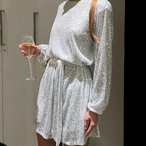 May include: A silver sequined mini dress with long sleeves and a tie belt. The dress has a scoop neckline and a relaxed fit.