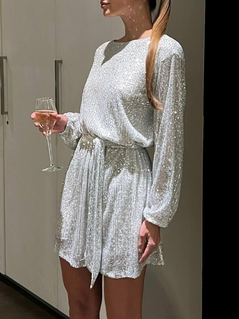May include: A silver sequined mini dress with long sleeves and a tie belt. The dress has a scoop neckline and a relaxed fit.
