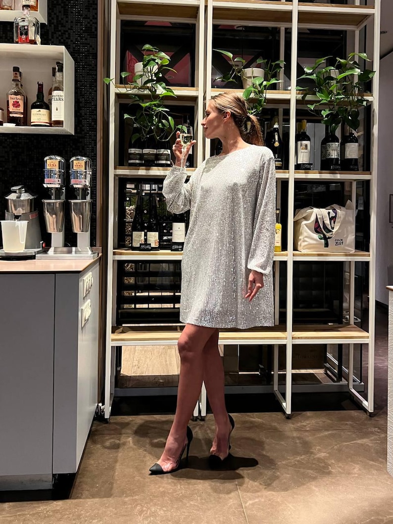 May include: A woman wearing a silver sequined dress and black heels stands in front of a white shelving unit. The shelves are filled with various items, including plants, bottles, and a white tote bag with the text "LOVE 2017" printed on it.