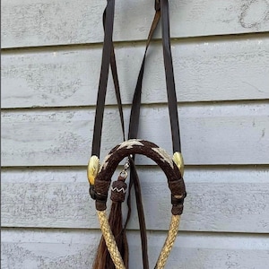 Puede incluir: Un cabezal de caballo marrón y beige con detalles dorados. El cabezal presenta una muserola trenzada, una borla decorativa y correas de cuero. El cabezal está diseñado para uso ecuestre y cuelga contra una pared de madera desgastada.