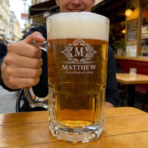 May include: A large, clear glass beer mug filled with golden beer and a foamy head. The mug has a handle and is engraved with the name "MATTHEW" and "Established 1986" along with a decorative initial "M".