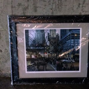 May include: A framed print of a city scene with tall buildings, trees, and a horse-drawn carriage. The print is in a black frame and is covered in plastic wrap.