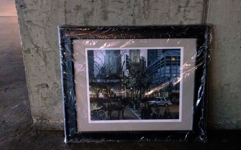 May include: A framed print of a city scene with tall buildings, trees, and a horse-drawn carriage. The print is in a black frame and is covered in plastic wrap.