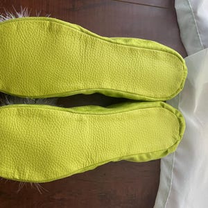 May include: A pair of lime green slippers with a textured, pebbled surface on the soles. The slippers feature a rounded toe design and are accented with soft, light-colored feathers extending from the front edges of the footwear.