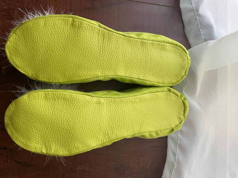May include: A pair of lime green slippers with a textured, pebbled surface on the soles. The slippers feature a rounded toe design and are accented with soft, light-colored feathers extending from the front edges of the footwear.