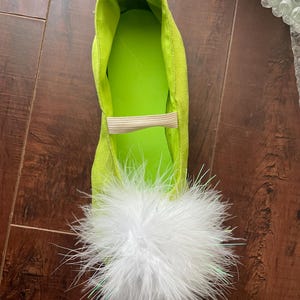 May include: A bright lime green flat shoe featuring a textured exterior, a smooth lime green interior lining, and a beige elastic strap across the top. The toe area is adorned with a large, voluminous white feather pom-pom decoration.