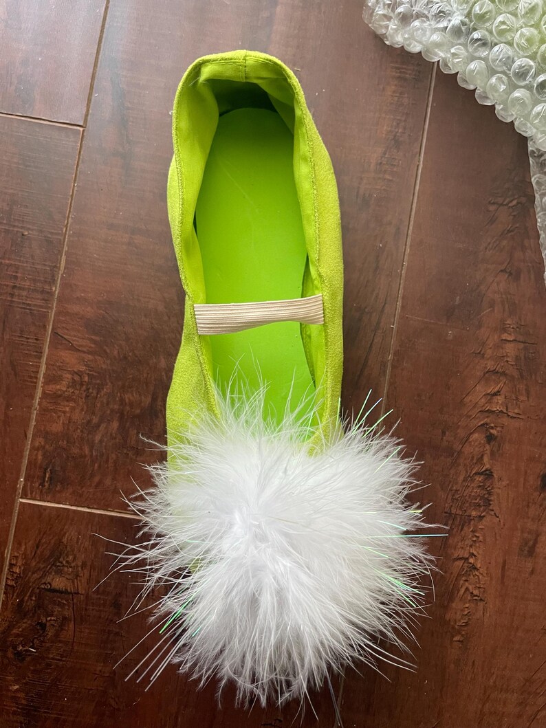 May include: A bright lime green flat shoe featuring a textured exterior, a smooth lime green interior lining, and a beige elastic strap across the top. The toe area is adorned with a large, voluminous white feather pom-pom decoration.