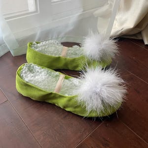 May include: A pair of lime green slip-on flats featuring a soft, suede-like texture and a large, fluffy white feather pom-pom attached to the rounded toe. Each shoe includes a thin, light-colored elastic strap across the top and clear bubble wrap.
