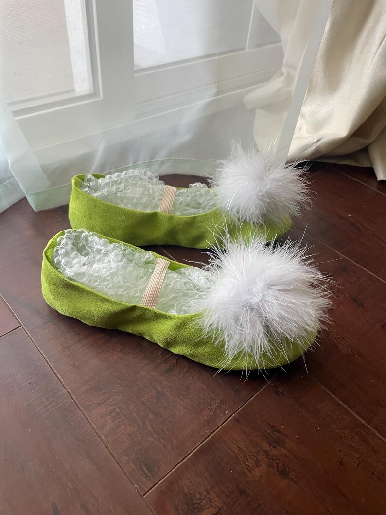 May include: A pair of lime green slip-on flats featuring a soft, suede-like texture and a large, fluffy white feather pom-pom attached to the rounded toe. Each shoe includes a thin, light-colored elastic strap across the top and clear bubble wrap.