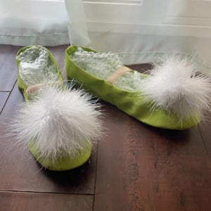 May include: A pair of lime green ballet flats featuring large, fluffy white pom-poms on the rounded toes. The shoes have a soft, fabric-like texture and include a thin elastic strap across the top. Clear bubble wrap is visible inside the shoes.