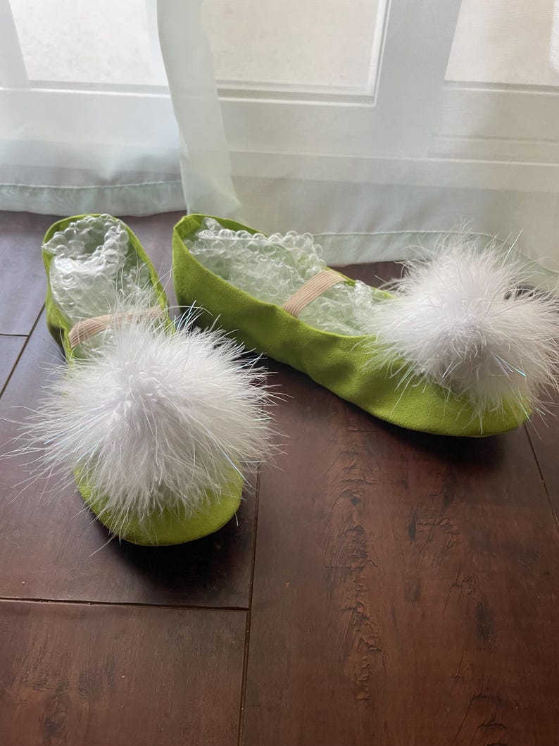May include: A pair of lime green ballet flats featuring large, fluffy white pom-poms on the rounded toes. The shoes have a soft, fabric-like texture and include a thin elastic strap across the top. Clear bubble wrap is visible inside the shoes.