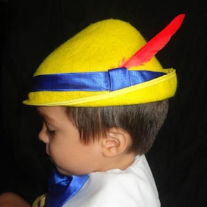 May include: A yellow felt hat with a blue ribbon and a red feather. The hat is a costume accessory, likely for a child's outfit. The hat has a curved brim and a dark background.