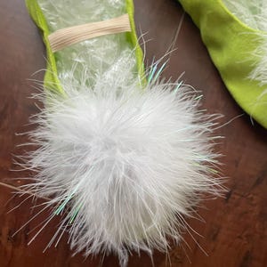 May include: A pair of lime green ballet slippers featuring a large, fluffy white pom-pom on the top of each shoe. The slippers are constructed from a soft, suede-like material and include a thin, beige elastic strap across the mid-foot area.