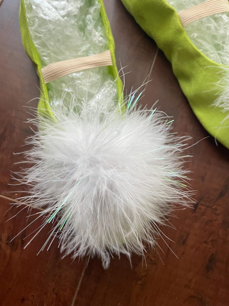 May include: A pair of lime green ballet slippers featuring a large, fluffy white pom-pom on the top of each shoe. The slippers are constructed from a soft, suede-like material and include a thin, beige elastic strap across the mid-foot area.