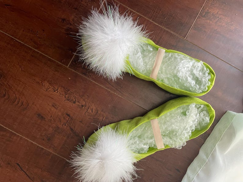 May include: A pair of lime green ballet slippers featuring a fluffy white feather pom-pom on the toe and a thin elastic strap across the midfoot. The interior of the slippers is filled with clear bubble wrap for shape retention.