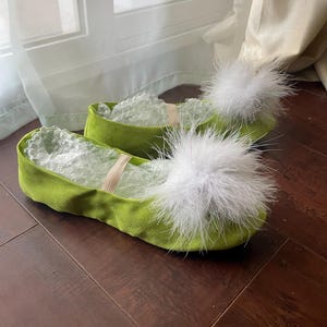 K&ouml;nnte beinhalten: Ein Paar lindgr&uuml;ne Slipper mit abgerundeter Spitze und einem gro&szlig;en, flauschigen wei&szlig;en Feder-Pompon auf der Oberseite jedes Schuhs. Die Schuhe haben eine weiche, wildlederartige Textur und verf&uuml;gen &uuml;ber einen d&uuml;nnen, elastischen Riemen.