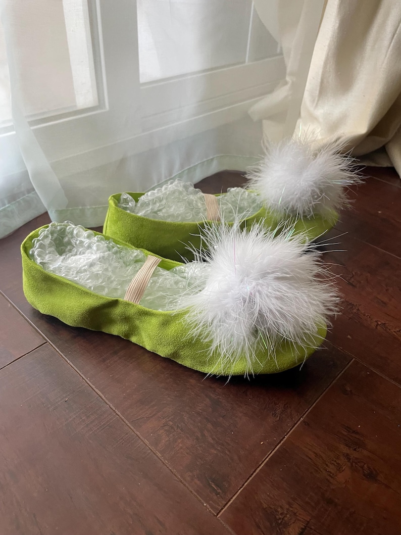 K&ouml;nnte beinhalten: Ein Paar lindgr&uuml;ne Slipper mit abgerundeter Spitze und einem gro&szlig;en, flauschigen wei&szlig;en Feder-Pompon auf der Oberseite jedes Schuhs. Die Schuhe haben eine weiche, wildlederartige Textur und verf&uuml;gen &uuml;ber einen d&uuml;nnen, elastischen Riemen.