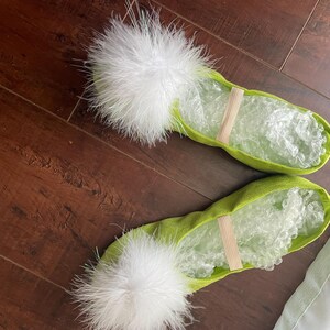 K&ouml;nnte beinhalten: Ein Paar lindgr&uuml;ne Ballettschl&auml;ppchen mit einem flauschigen wei&szlig;en Feder-Pompon an der Spitze und einem d&uuml;nnen elastischen Riemen &uuml;ber dem Mittelfu&szlig;. Das Innere der Schl&auml;ppchen ist zur Formgebung mit transparenter Luftpolsterfolie gef&uuml;llt.