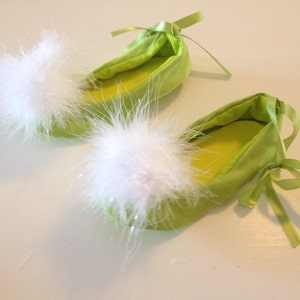 May include: A pair of lime green ballet-style slippers featuring a soft, white, fluffy pom-pom on the rounded toe of each shoe. The slippers are accented with matching lime green satin ribbon ties at the heel and a smooth, solid-colored interior.