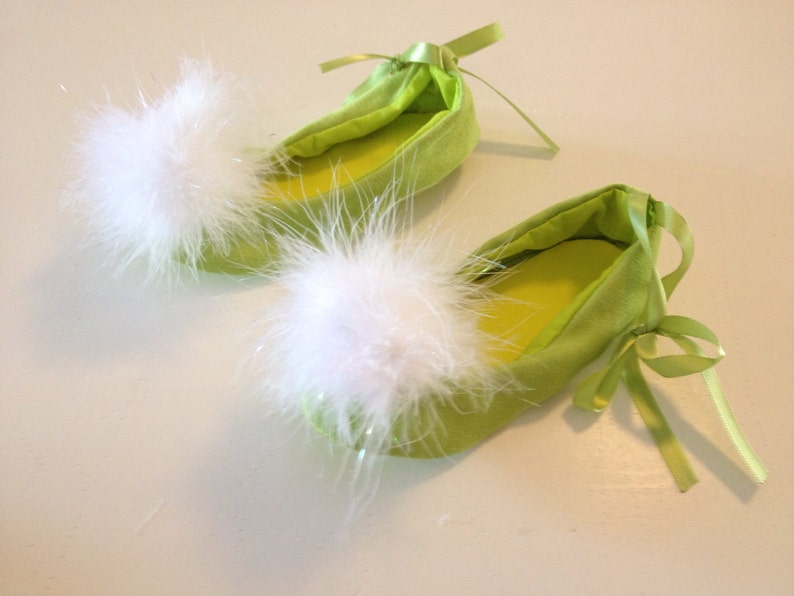 May include: A pair of lime green ballet-style slippers featuring a soft, white, fluffy pom-pom on the rounded toe of each shoe. The slippers are accented with matching lime green satin ribbon ties at the heel and a smooth, solid-colored interior.
