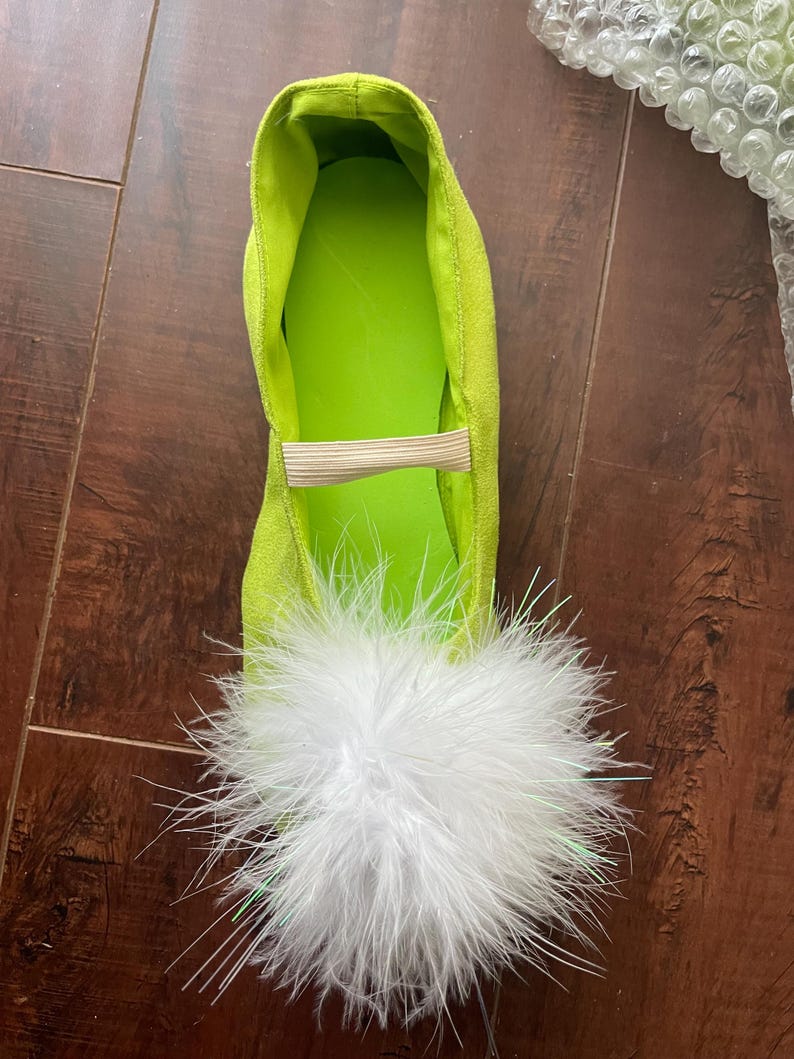 K&ouml;nnte beinhalten: Ein flacher Schuh in hellem Limettengr&uuml;n mit strukturierter Au&szlig;enseite, glattem limettengr&uuml;nen Innenfutter und einem beigen elastischen Riemen &uuml;ber dem Spann. Der Zehenbereich ist mit einem gro&szlig;en, volumin&ouml;sen wei&szlig;en Feder-Pompon verziert.