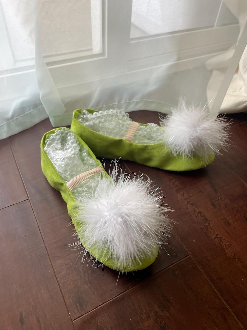 K&ouml;nnte beinhalten: Ein Paar lindgr&uuml;ne Ballettschl&auml;ppchen mit abgerundeter Spitze und einem gro&szlig;en, flauschigen wei&szlig;en Bommel auf der Oberseite. Jeder Schuh verf&uuml;gt &uuml;ber ein d&uuml;nnes, beiges Gummiband &uuml;ber dem Spann und ist mit transparenter Luftpolsterfolie ausgef&uuml;llt.