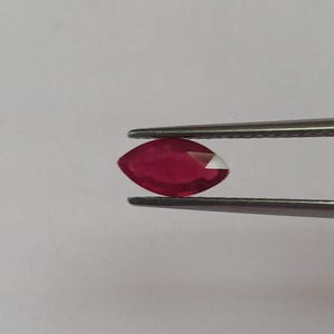 May include: A marquise-cut ruby gemstone, held by tweezers, against a white background. The ruby is a deep red colour with a faceted surface and a white tip. The tweezers are silver.