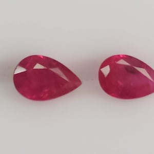 May include: Two faceted, pear-shaped rubies. The gemstones are a deep, vibrant red color, with multiple facets catching the light. The rubies are set against a plain white background.
