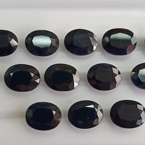May include: Twelve oval-cut, black gemstones are arranged in three rows on a white surface. The gems have a polished, reflective surface, with facets catching the light. The gemstones are of similar size and shape, presented in a neat arrangement.
